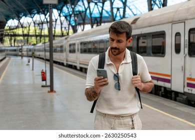 Young man with a backpack checking a mobile app at the railway station, waiting for his train while planning his next journey
