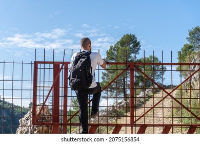 Young Man With Backpack Climbing Through A Gate With Metal Fence.