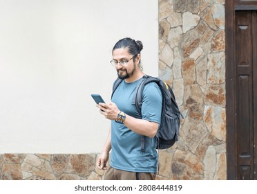 Young man with a backpack looking down at his mobile phone screen while walking past a colonial stone wall.