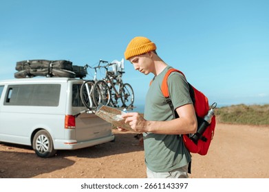 Young man with a backpack looking at the map, near a van with mountain bikes ready to start the biking 