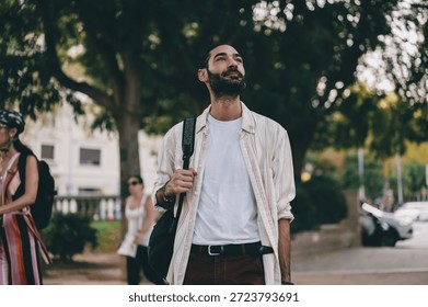 Young man with backpack looks up while walking in city park, wearing striped shirt and white tee, expressing inspiration, curiosity and urban freedom with calm presence.