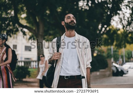 Young man with backpack looks up while walking in city park, wearing striped shirt and white tee, expressing inspiration, curiosity and urban freedom with calm presence.