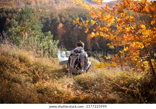 a young man\
with a backpack and a map is sitting on the bank of a cliff by the\
river in autumn, traveling\
alone