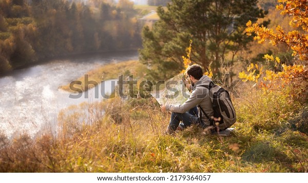a young man\
with a backpack and a map is sitting on the bank of a cliff by the\
river in autumn, traveling\
alone