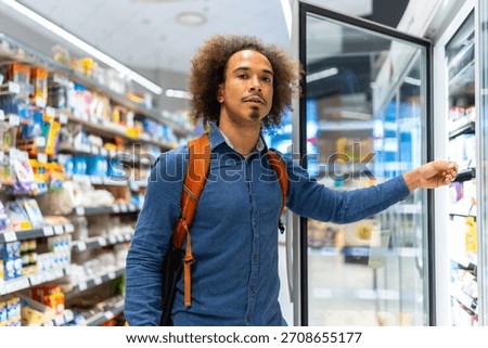 Young man with backpack selecting chilled items from a refrigerated display in a bright modern supermarket aisle, thoughtful consumer choosing fresh grab and go options