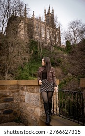 A young man with a backpack smiling at the camera while standing on a bridge over the Water of Leith, with historic buildings in the background.