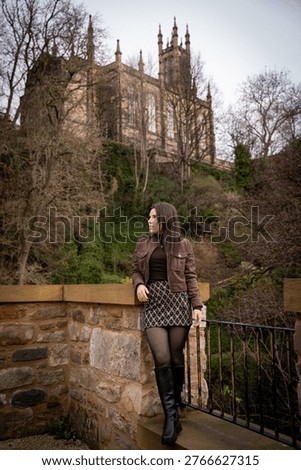 A young man with a backpack smiling at the camera while standing on a bridge over the Water of Leith, with historic buildings in the background.