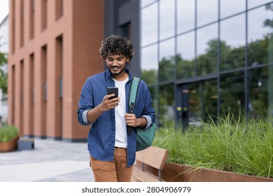 Young man with a backpack smiling while confidently walking and checking his mobile phone outside a contemporary building, connecting with technology and the urban environment