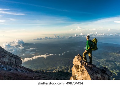 Young man with backpack standing on a cliffs edge on the top of mountain with gorgeous view to forests, clouds and sea. Volcano Agung, Bali, Indonesia.	

