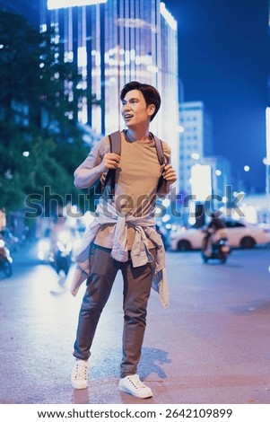A young man with a backpack strolls through vibrant downtown Hanoi at night, illuminated by bright lights and surrounded by busy streets and vehicles.