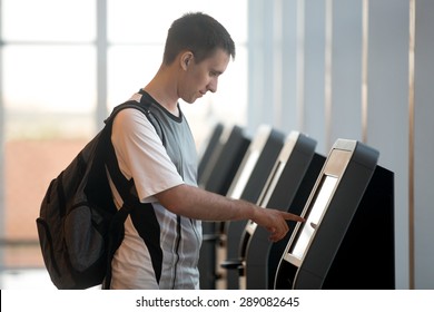 Young man with backpack touching interactive display at self-service transfer machine, doing self-check-in for flight or buying airplane tickets at automatic device in modern airport terminal building