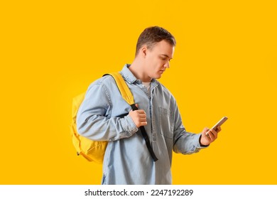 Young man with backpack using mobile phone on yellow background