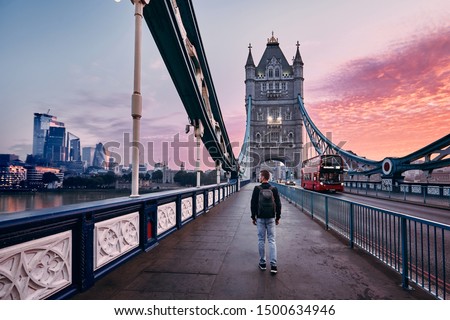 Young man with backpack walking on Tower Bridge against cityscape with skyscrapes at colorful sunrise. London, United Kingdom