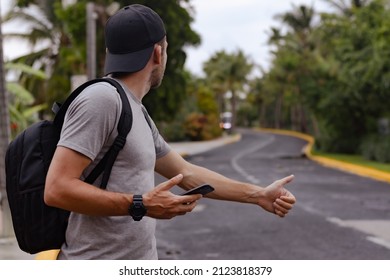 Young Man Backpacker In Cap Stands On The Road With Smartphone And Thumb Up And Hitchhiking. Man Holds Mobile Phone In Hand And Catches A Taxi.