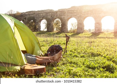 Young man backpacker sleeping in camping tent on grass with feet outside in front of roman aqueduct ruins in park in rome at sunset. Walking sticks and guitar.