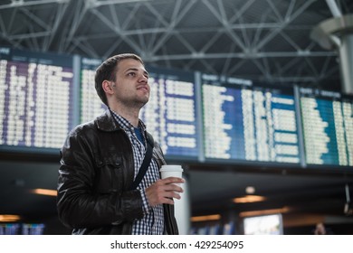 Young man with a bag in airport near flight timetable holding cup of coffee