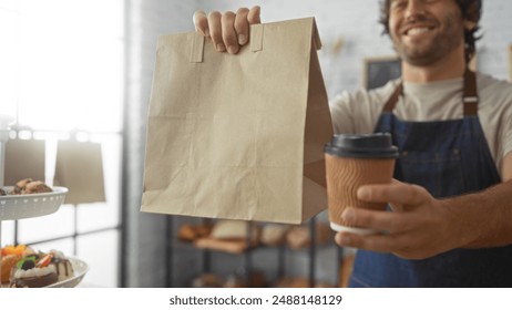 Young man in bakery holding a paper bag and coffee cup with pastries displayed in the background - Powered by Shutterstock