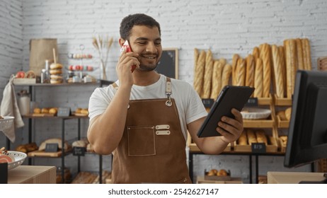 Young man in a bakery shop talking on the phone while holding a tablet, showing an attractive and happy worker in an indoor setting with bread in the background. - Powered by Shutterstock