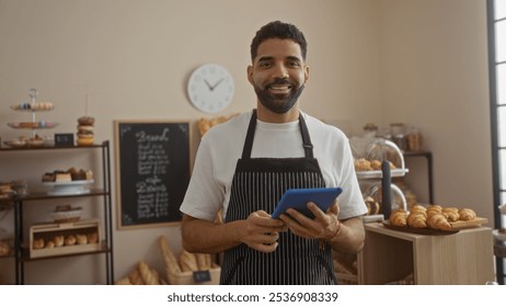 Young man in bakery with tablet smiling, surrounded by shelves of bread, pastries, macaroons, and a blackboard menu, standing in front of a clock and window, wearing a striped apron. - Powered by Shutterstock