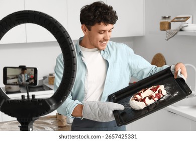 Young man baking bread for online cooking class, recording video with smartphone and ring light in modern kitchen, he holds a baking tray with raw dough topped with fruit and nuts