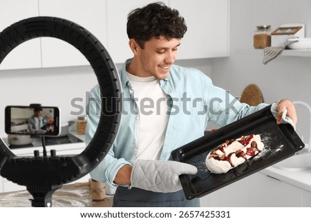 Young man baking bread for online cooking class, recording video with smartphone and ring light in modern kitchen, he holds a baking tray with raw dough topped with fruit and nuts