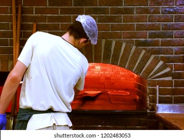 Young Man Baking A Pizza In A Professional Brick Oven With Red Light Due To The Fire Inside The Oven