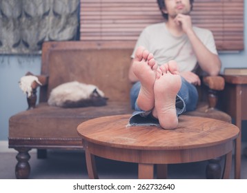 A Young Man With Bare Feet Is Resting On A Sofa At Home With A Cat Sleeping Next To Him