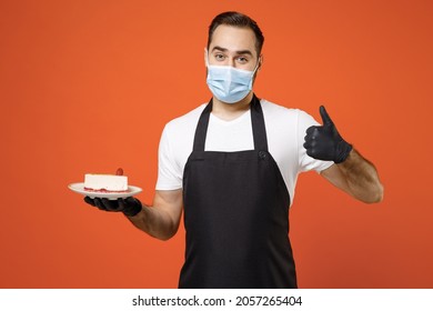 Young Man Barista Barman Employee In Apron White T-shirt Sterile Face Mask Coronavirus Covid19 Pandemic Quarantine Work Coffee Shop Giving Cake Dessert Isolated On Orange Background Business Startup