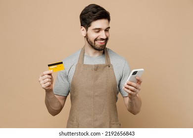 Young Man Barista Barman Employee Wear Brown Apron Work In Coffee Shop Using Mobile Cell Phone Credit Bank Card Shopping Online Isolated On Plain Light Beige Background Small Business Startup Concept