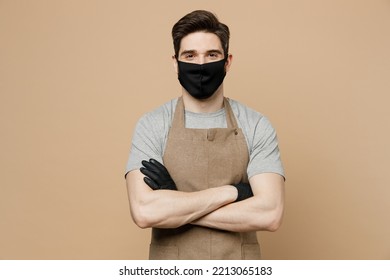 Young Man Barista Barman Employee Wear Brown Apron Black Sterile Mask Glowes Hold Hand Crossed Folded Work In Coffee Shop Isolated On Plain Pastel Light Beige Background Small Business Startup Concept