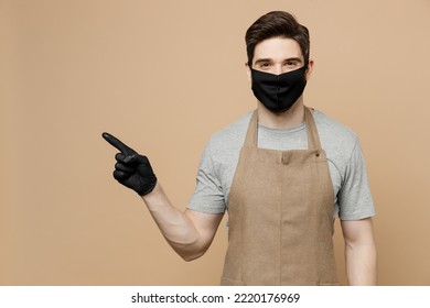 Young Man Barista Barman Employee Wear Brown Apron Black Sterile Mask Point Finger Aside On Workspace Work In Coffee Shop Isolated On Plain Pastel Light Beige Background Small Business Startup Concept