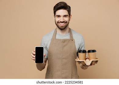 Young Man Barista Barman Employee Wear Brown Apron Work In Shop Hold Craft Cup Coffee To Go Blank Screen Mobile Phone Isolated On Plain Pastel Light Beige Background. Small Business Startup Concept