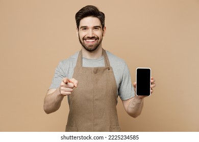 Young Man Barista Barman Employee Wear Brown Apron Work In Coffee Shop Hold Blank Screen Mobile Cell Phone Point Finger Camera On You Isolated On Plain Beige Background Small Business Startup Concept