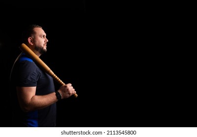 Young Man With A Baseball Bat On A Dark Background. Bouncer, Security Guard, Bodyguard Concept. Place For Text. A Large Man In A T-shirt Stands Sideways.