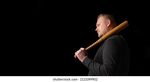 Young Man With A Baseball Bat On A Dark Background. Bouncer, Security Guard, Bodyguard Concept. Place For Text. A Large Man In A T-shirt Stands Sideways.