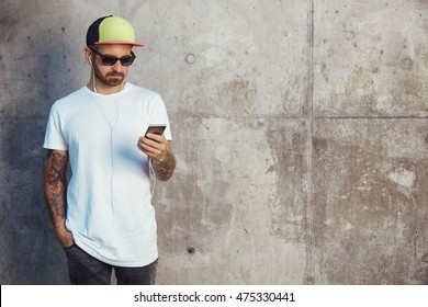 Young man in baseball cap, sunglasses and white blank t-shirt reading something on his smartphone standing next to a gray concrete wall - Powered by Shutterstock