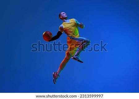 Young man, basketball player in yellow uniform training, throwing ball in a jump against blue studio background in neon light. Concept of professional sport, hobby, healthy lifestyle, action, motion
