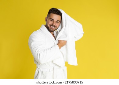 Young man in bathrobe drying hair with towel on yellow background