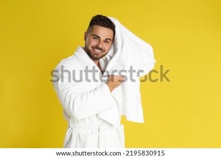 Young man in bathrobe drying hair with towel on yellow background
