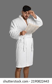 Young Man In Bathrobe Reading Newspaper On Grey Background