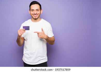 Young Man With A Beaming Smile And Pointing To A New Credit Card. Cheerful Man Holding A Credit Card In A Purple Background With Copy Space