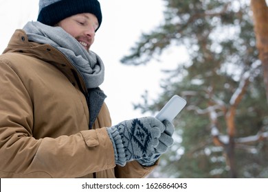 Young man in beanie, scarf, jacket and gloves scrolling in smartphone on winter day in snowbound forest