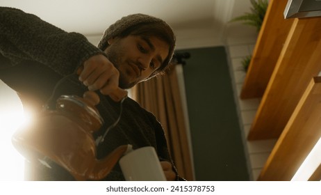 Young man with beanie stands in the modern kitchen, pours tea from a teapot into a mug in his hand, leans against the kitchen counter and drinks his tea in the morning. Low angle shot - Powered by Shutterstock