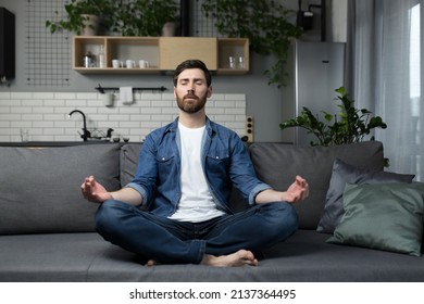 Young man with a beard alone at home, sitting on the couch and meditating in the lotus position, in casual clothes - Powered by Shutterstock