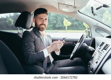 Young Man With A Beard Businessman Driving A Car In The Parking Lot Smiles And Writes On The Phone Reads The News Successful Man