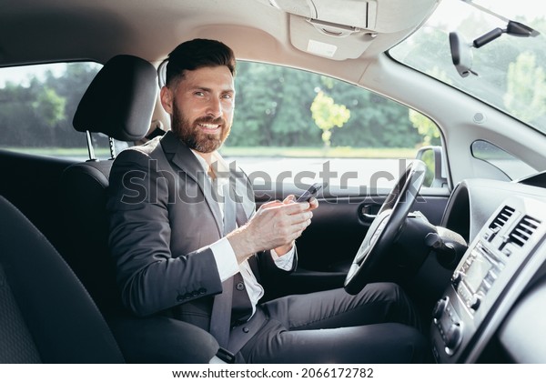 Young man with a beard businessman driving a car in\
the parking lot smiles and writes on the phone reads the news\
successful man