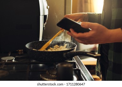 Young Man With Beard Cooking Dinner And Using His Smart Phone At The Kitchen.