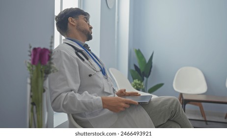 A young man with a beard, dressed as a doctor, sitting pensively in a modern hospital lobby, holding a tablet. - Powered by Shutterstock