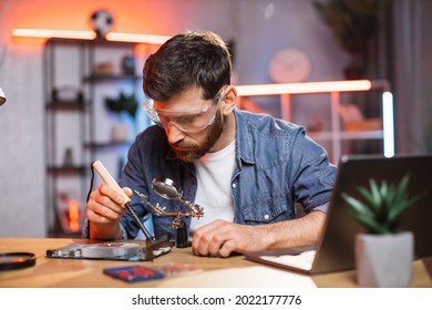 Young man with beard fixing display card from laptop using soldering iron. Handsome guy wearing protective glasses while repairing detail from computer. - Shutterstock ID 2022177776