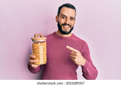 Young man with beard holding jar with macaroni pasta smiling happy pointing with hand and finger 